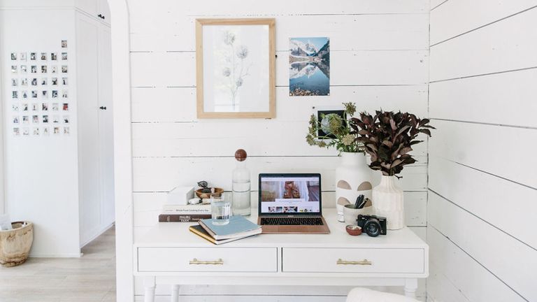 Bright and breezy home office set-up with white paneled walls, nature-inspired pictures on wall, and white desk with vases of foliage, laptop, and water carafe.