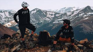 Two caucasian men wearing Fox mountain biking kit resting at the top of a mountain. One is sitting on the ground and looking up at the other who stands. They're both smiling.