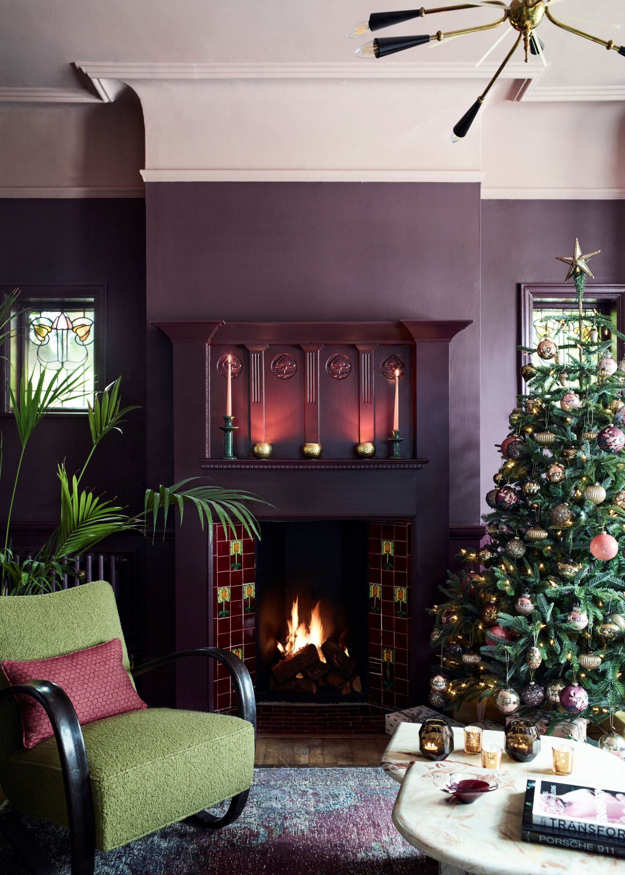 A deep plum--hued living room with pops of green decor is decorated for christmas with a large christmas tree adorned with plum and pink baubles and two taper candles are lit above the lit fireplace