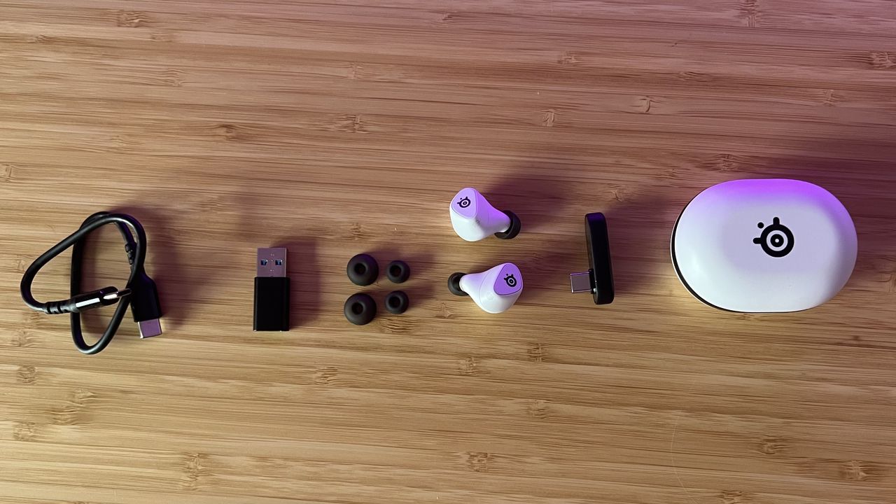 SteelSeries Arctis Game Buds with case, receiver, USB-A adapter, and cable on a wooden desk