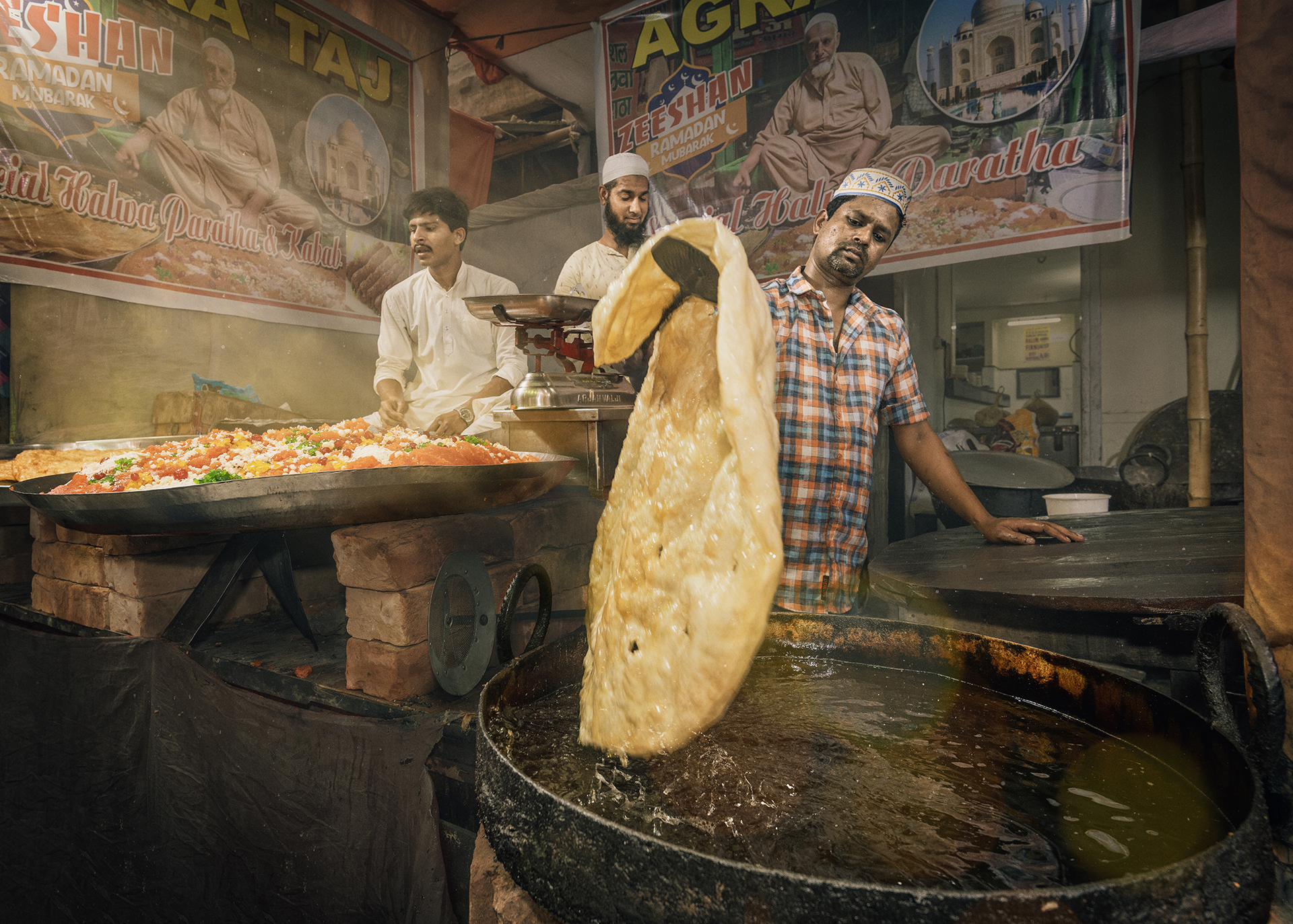 A vendor flips dough into hot oil at a bustling street food stall, with colorful dishes and festive banners in the background