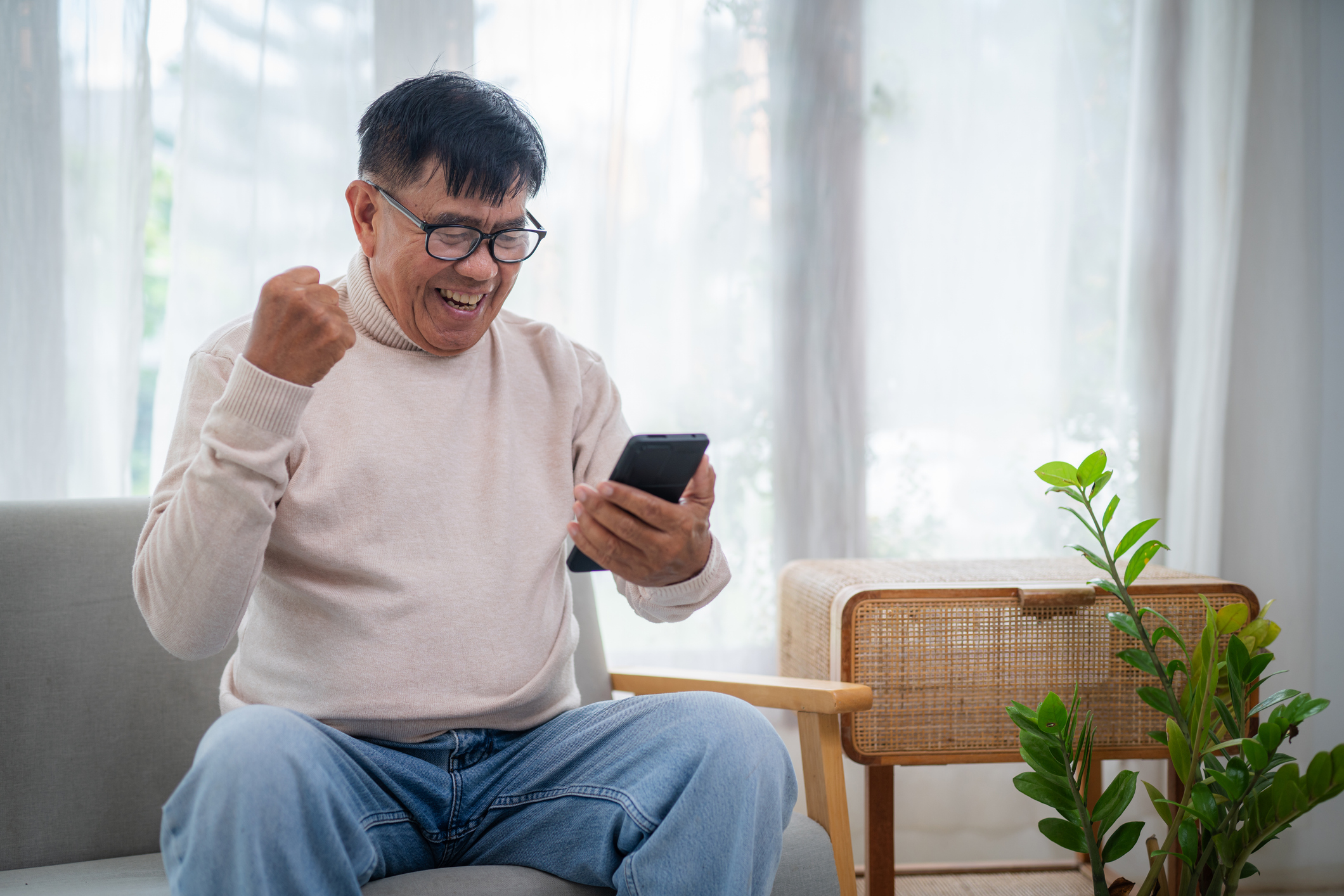 Asian happy older man sitting on comfortable sofa at home, using smartphone