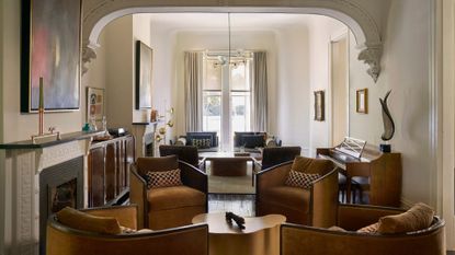 A bright white living room with brown armchairs, patterned pillows, arched ceiling, framed painting, stone mantel over the fireplace, and a large piano
