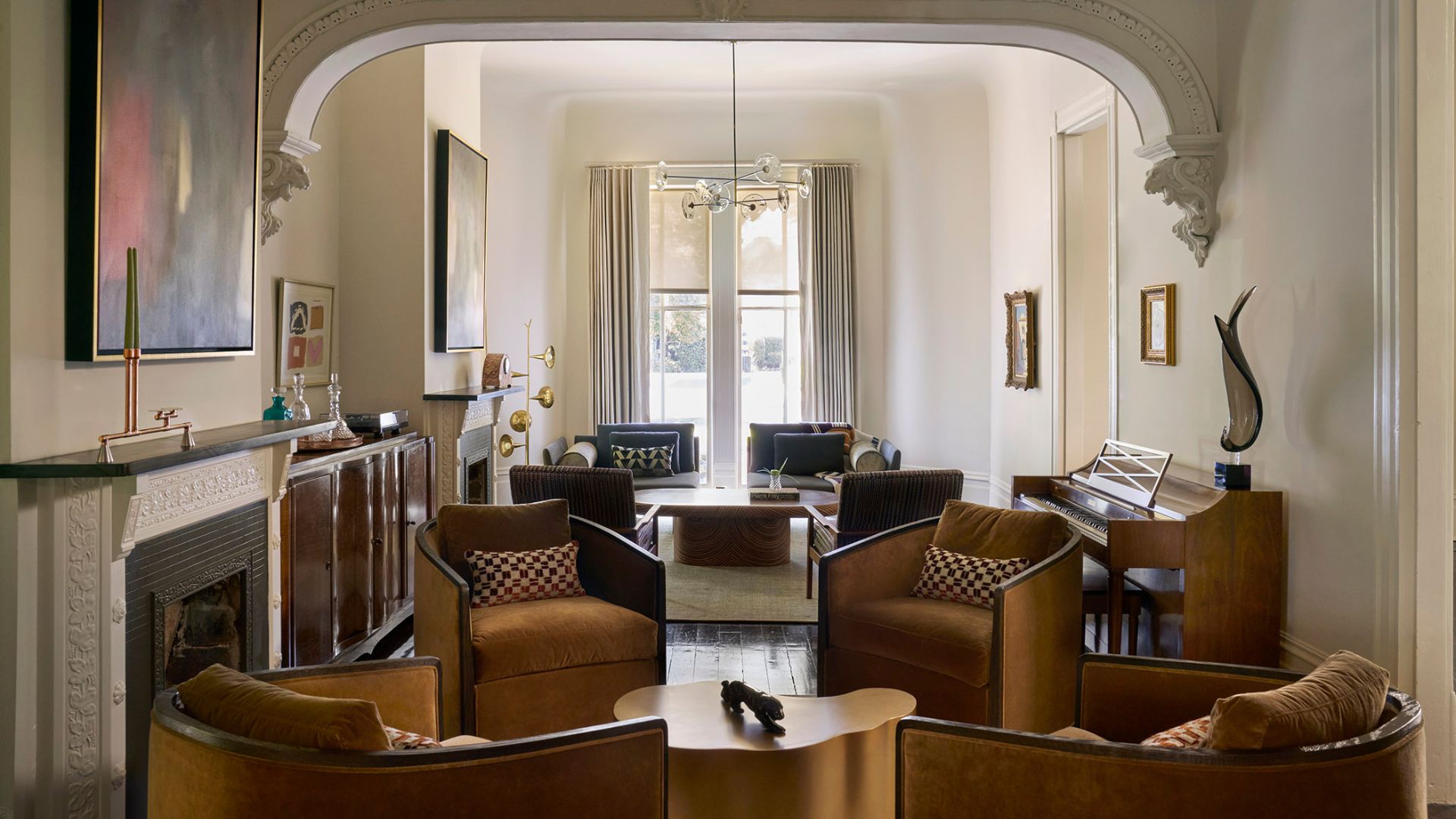 A bright white living room with brown armchairs, patterned pillows, arched ceiling, framed painting, stone mantel over the fireplace, and a large piano