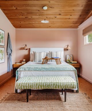 Mid-century style bench with green patterned padded seat at footend of bed, and breakfast tray on bed. Striped scatter pillows and green bed runner feature alongside rattan wall lights above bedsides on either side, and wood cladding on ceiling.