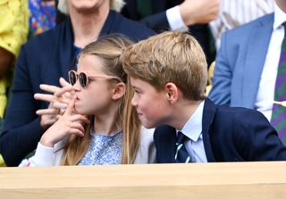 Princess Charlotte wearing sunglasses with her finger on her lip sitting next to Prince George at Wimbledon