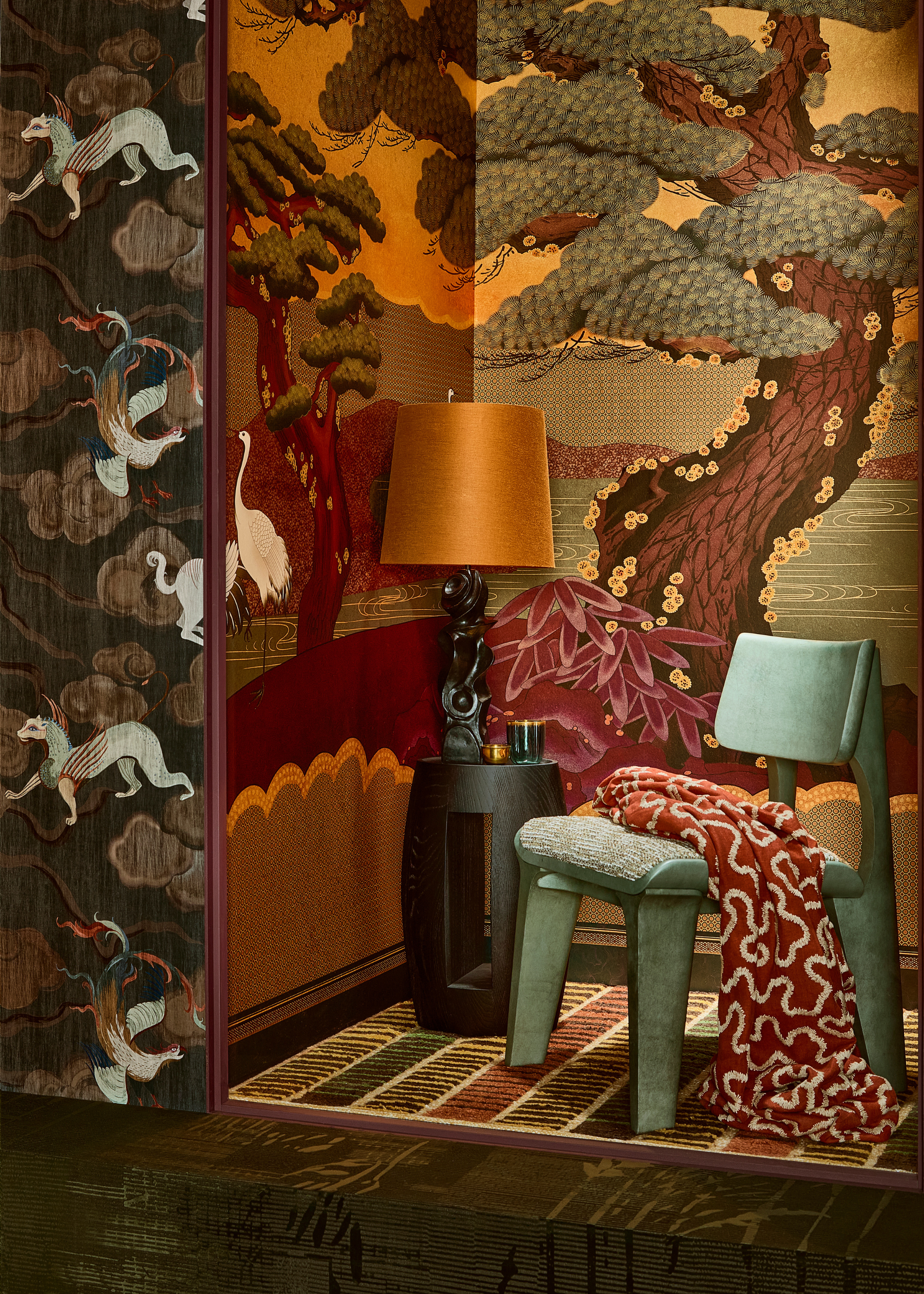 a design showcase featuring an alcove with a side table and table lamp, a green chair with a orange patterned throw over the seat with a small patterned rug on the floor and ornate japanese style wallpaper within the alcove and a different mythical creature ornate wallpaper on the walls outside of the alcove