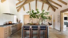 A large kitchen with tall ceilings featuring wooden beams, wooden cabinetry, gray countertops, a stainless steel island, and no upper cabinets of backsplash for a softer look
