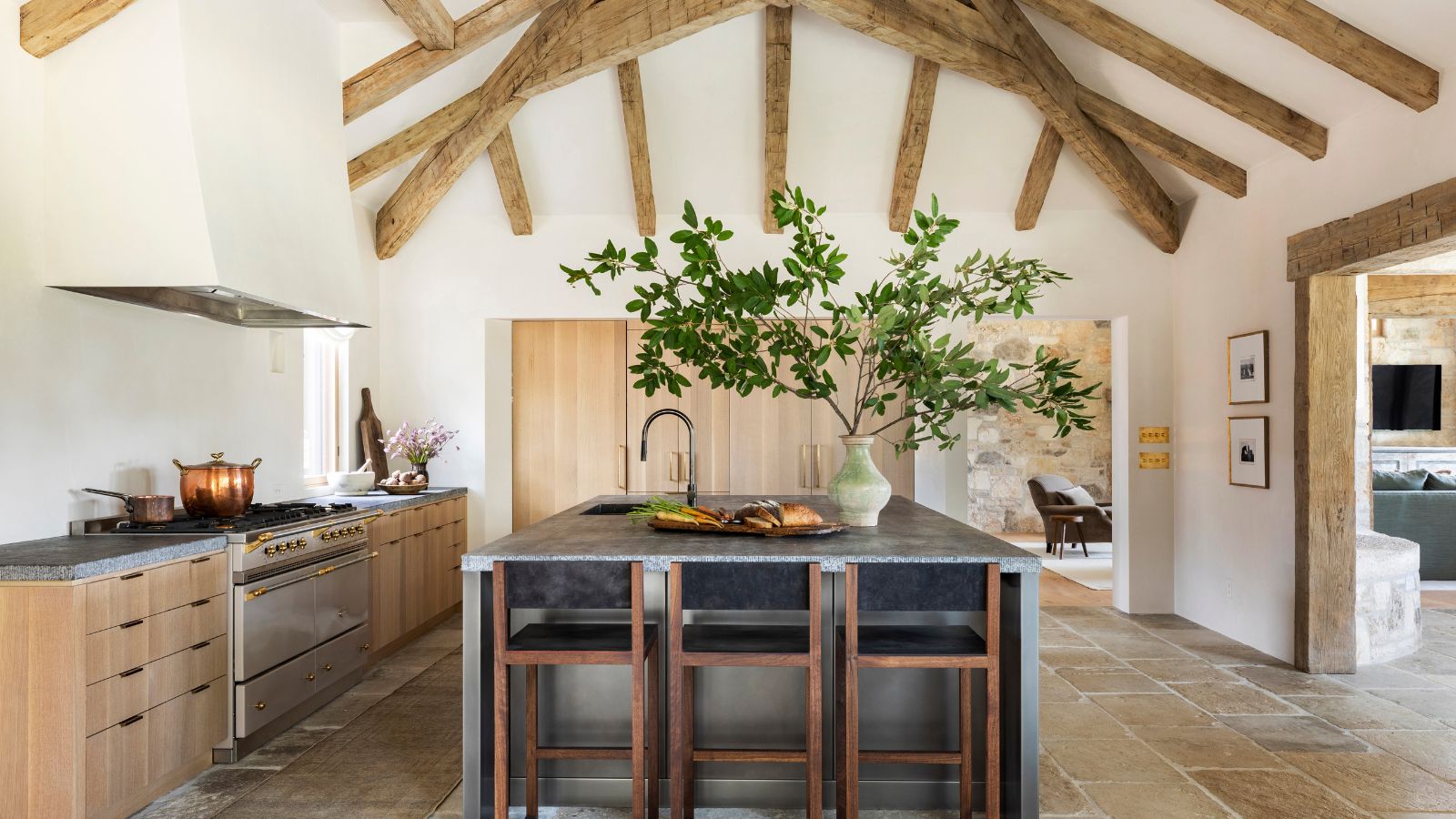 A large kitchen with tall ceilings featuring wooden beams, wooden cabinetry, gray countertops, a stainless steel island, and no upper cabinets of backsplash for a softer look