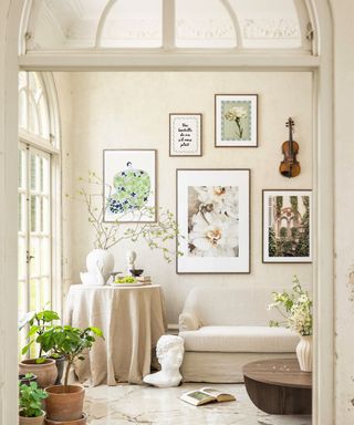 A beige living room with wall art on the walls, a side table with white decor on it, a beige couch, marble flooring with terracotta pots on it, and a glass arch above it