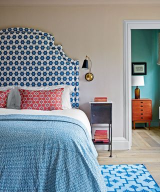 Bedroom with large, curvaceous blue and white patterned headboard, white and blue bedding with red cushions, metal wall lamp and bedside table, view into green room with table lamp and wooden table