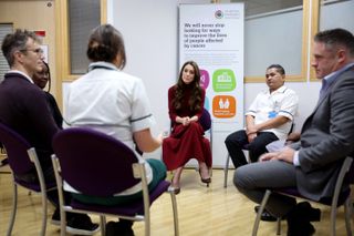 Kate Middleton wearing a wine skirt and turtleneck sitting in a chair in a circle around a group of other people at a cancer hospital