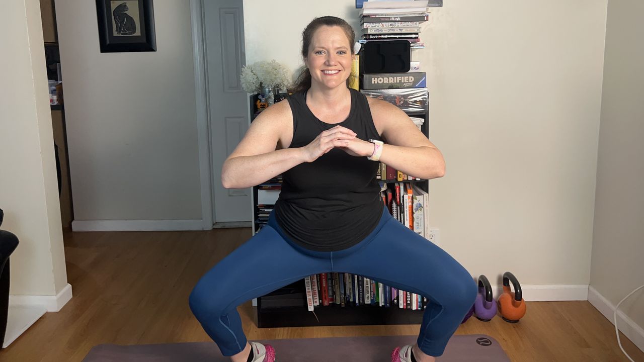 Trainer Jennifer Rizzuto performs a squat indoors. She is wearing leggings, a vest and sneakers and is exercising on a mat. Her knees are bent, pointing out to the side, and her hands are clasped at chest height. Behind her we see a picture on a wall, kettlebells and a bookcase.