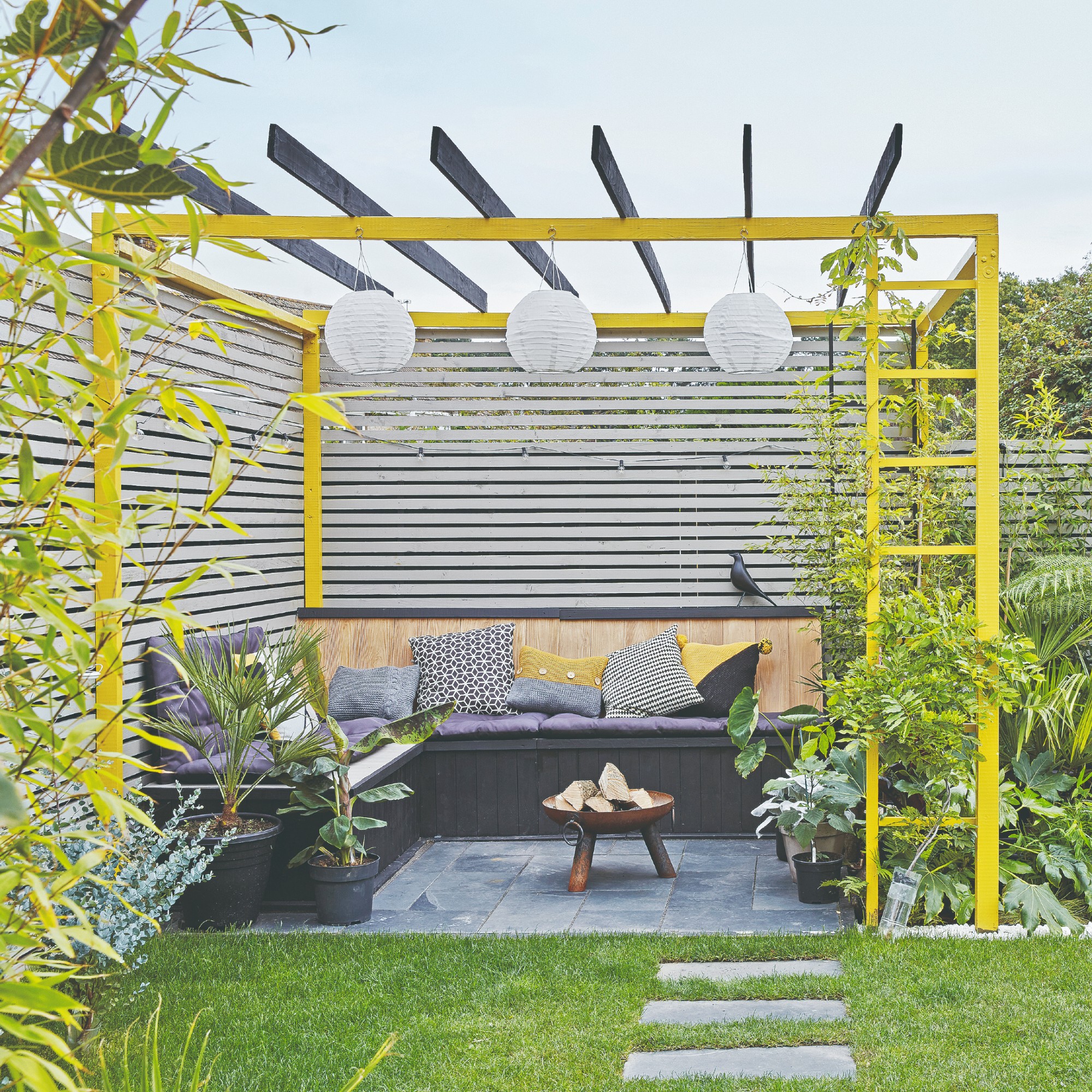 A garden with a built-in corner seating bench with cushions on top and a yellow pergola surrounding it