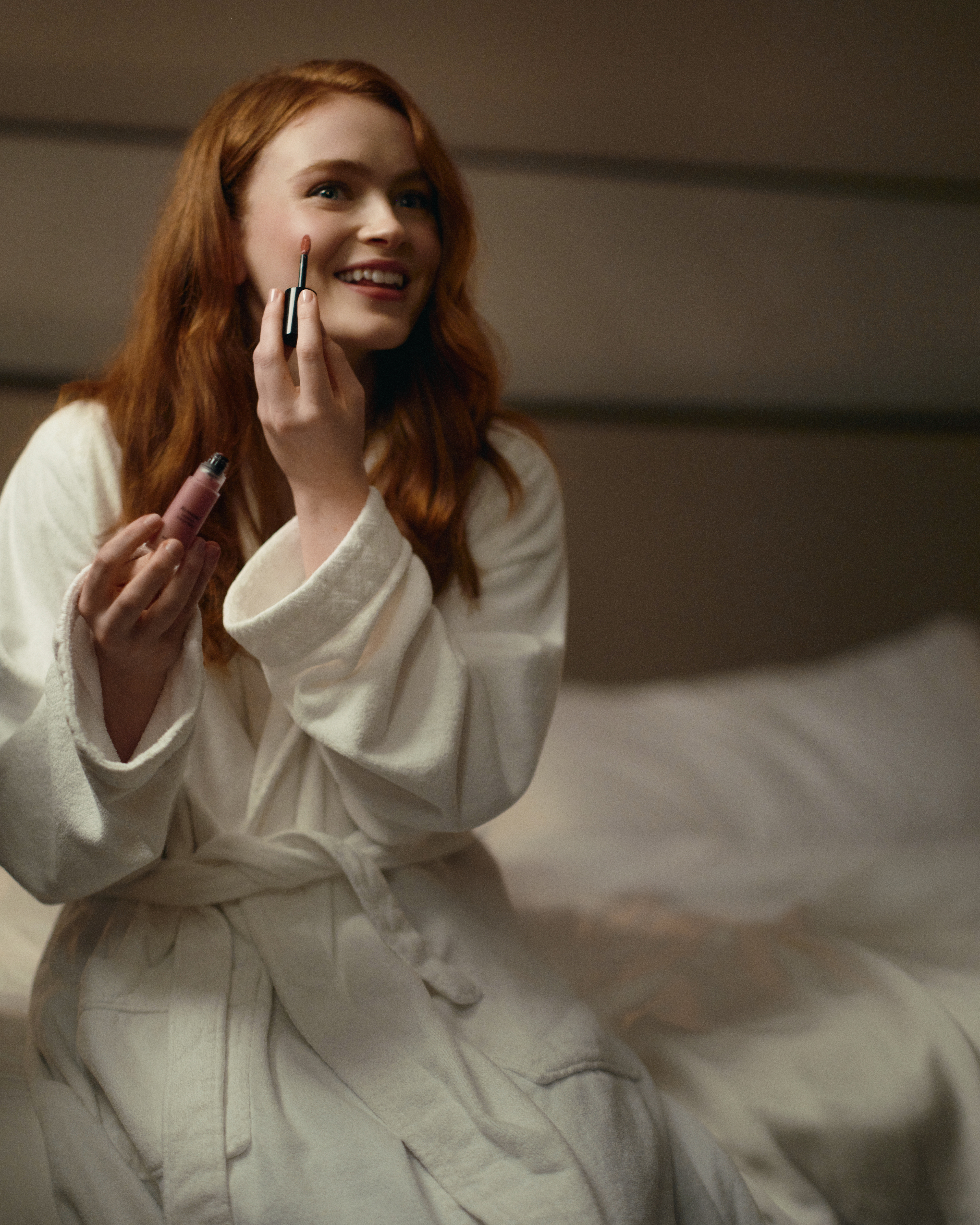 Sadie Sink wearing a belted robe and putting on Armani beauty products