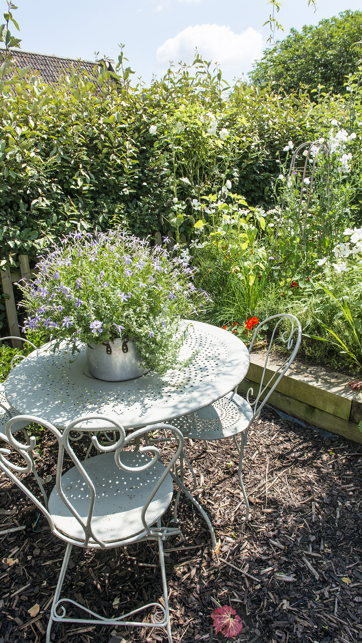 A small sheltered seating area with bark chippings on the ground, a round table and small metal chairs .