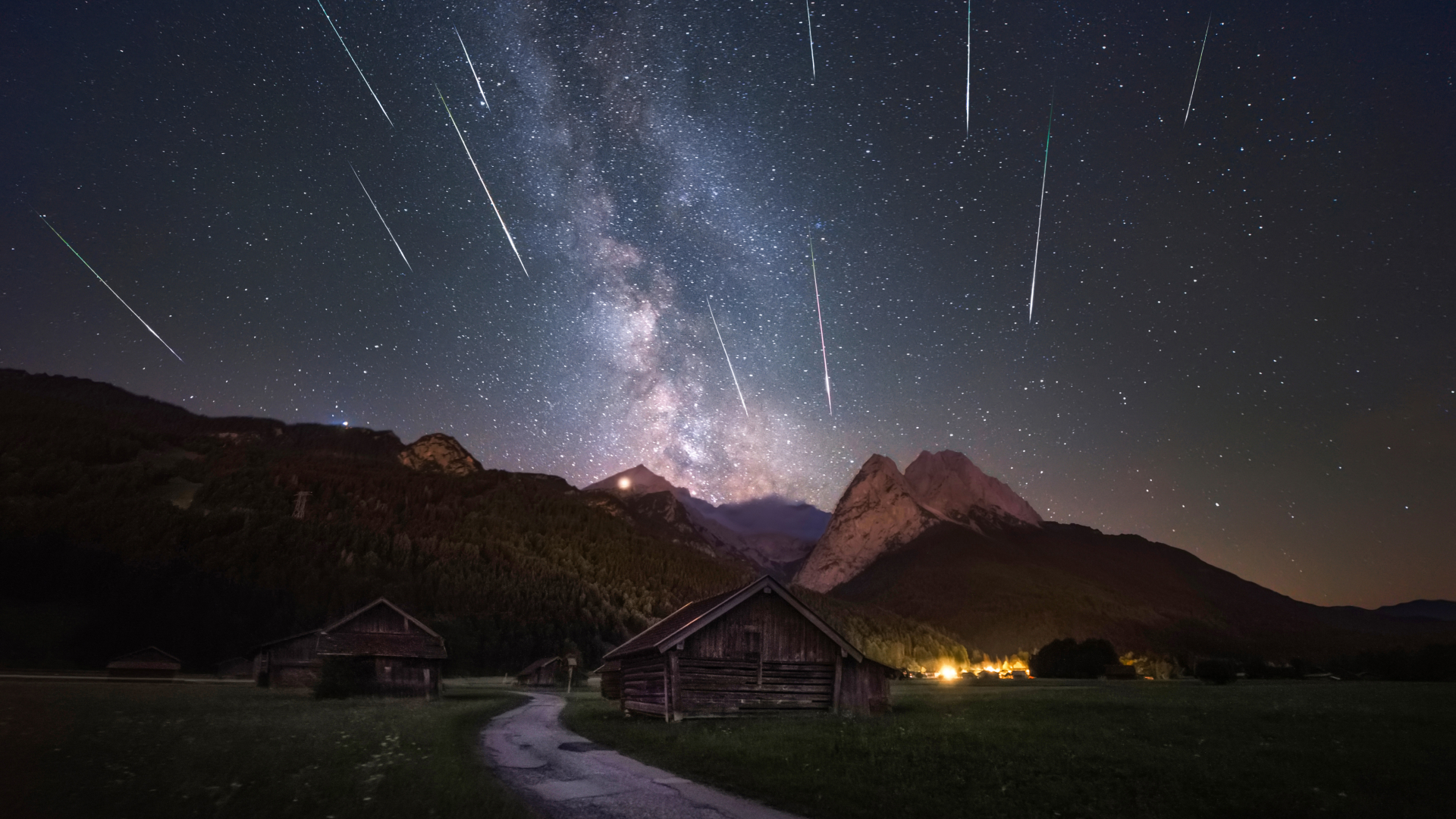 A cluster of bright shooting stars can be seen streaking through a night sky alongside the glowing band of the Milky Way above an alpine scene featuring two log cabins and a winding tarmac road.