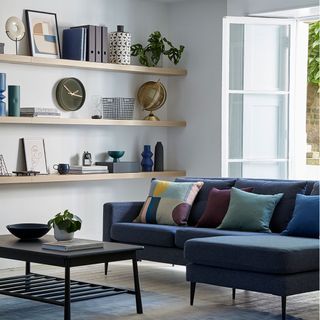 White living room with floating wall shelves, a blue sofa with colorful pillows and a coffee table