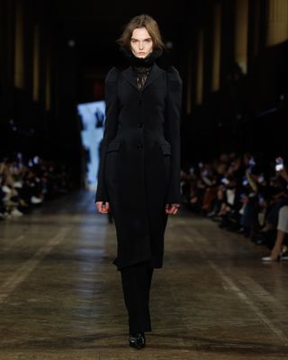 A model wearing a lace ruff blouse with a tailored coat and trousers walking at the F/W 25 Alexander McQueen show.