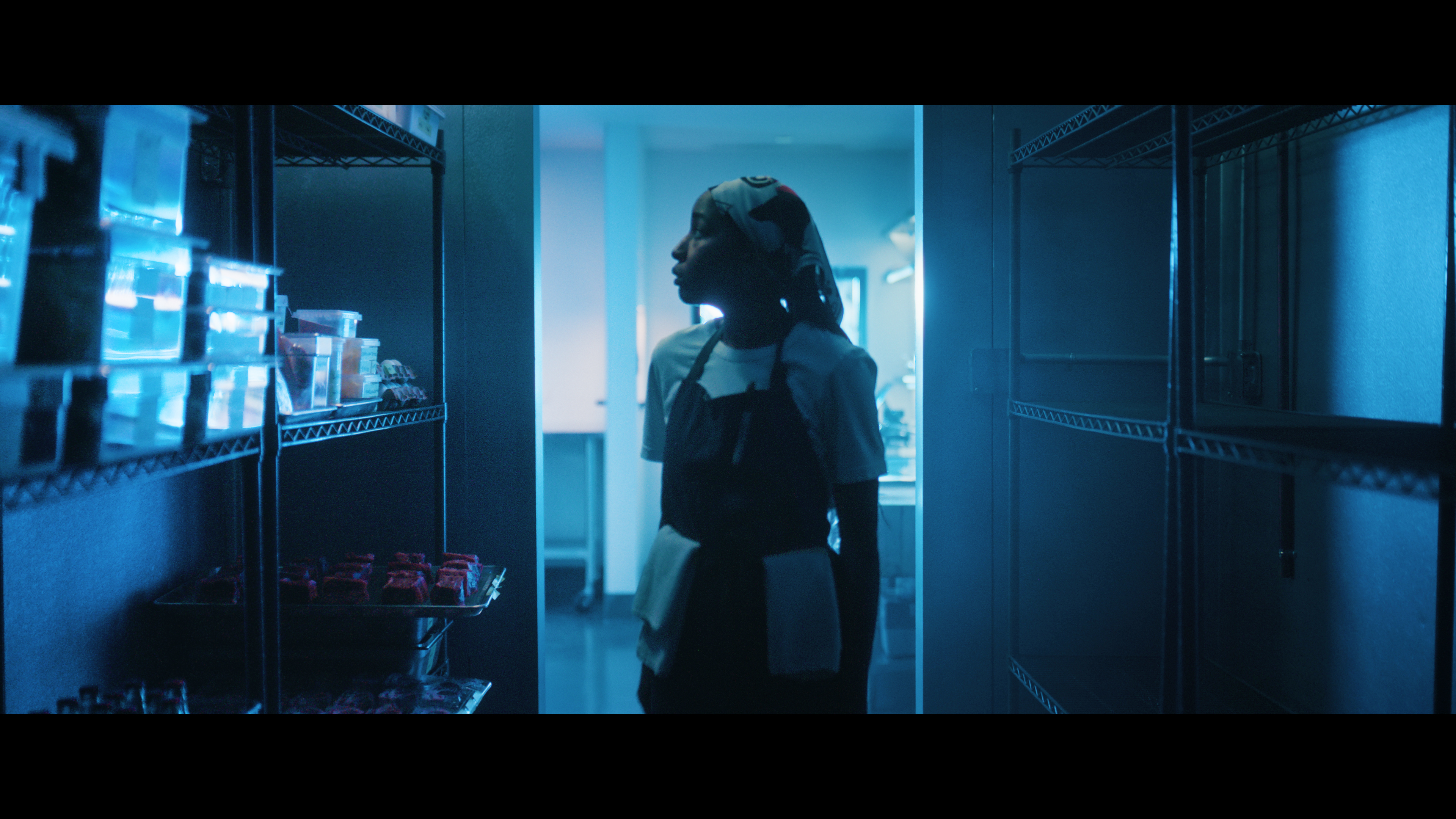 Ayo Edebiri as Sydney standing in a restaurant fridge and looking at a shelf of food with an apron on.