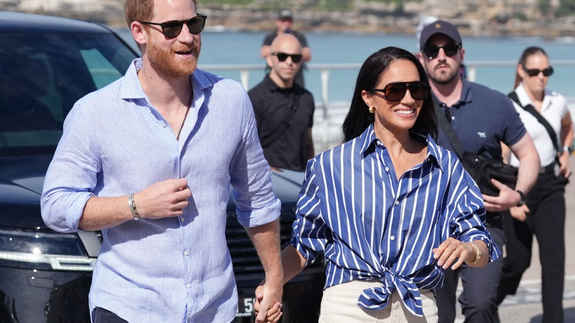 Meghan Markle holds hands with Prince Harry wearing white cropped jeans with white trainers and a blue striped shirt.
