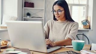 Dark-haired, smiling woman working from home with a silver laptop on a cluttered desk