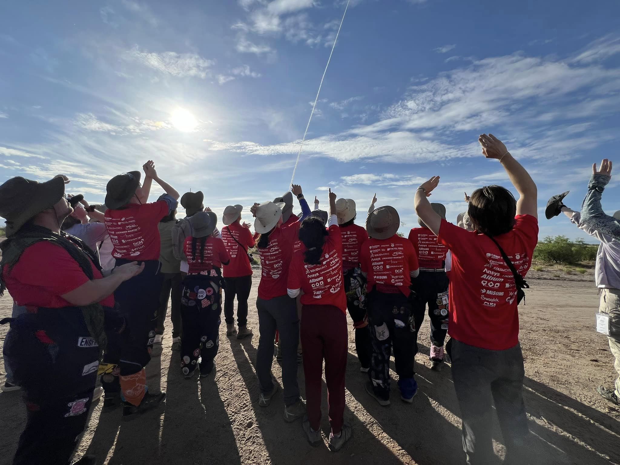 people in red shirts stare upwards as a rocket's smoke trail can be seen far in the distance