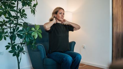 woman sitting on a velvet navy armchair looking off camera holding her neck. there's a plant to her side and a white wall behind her. 