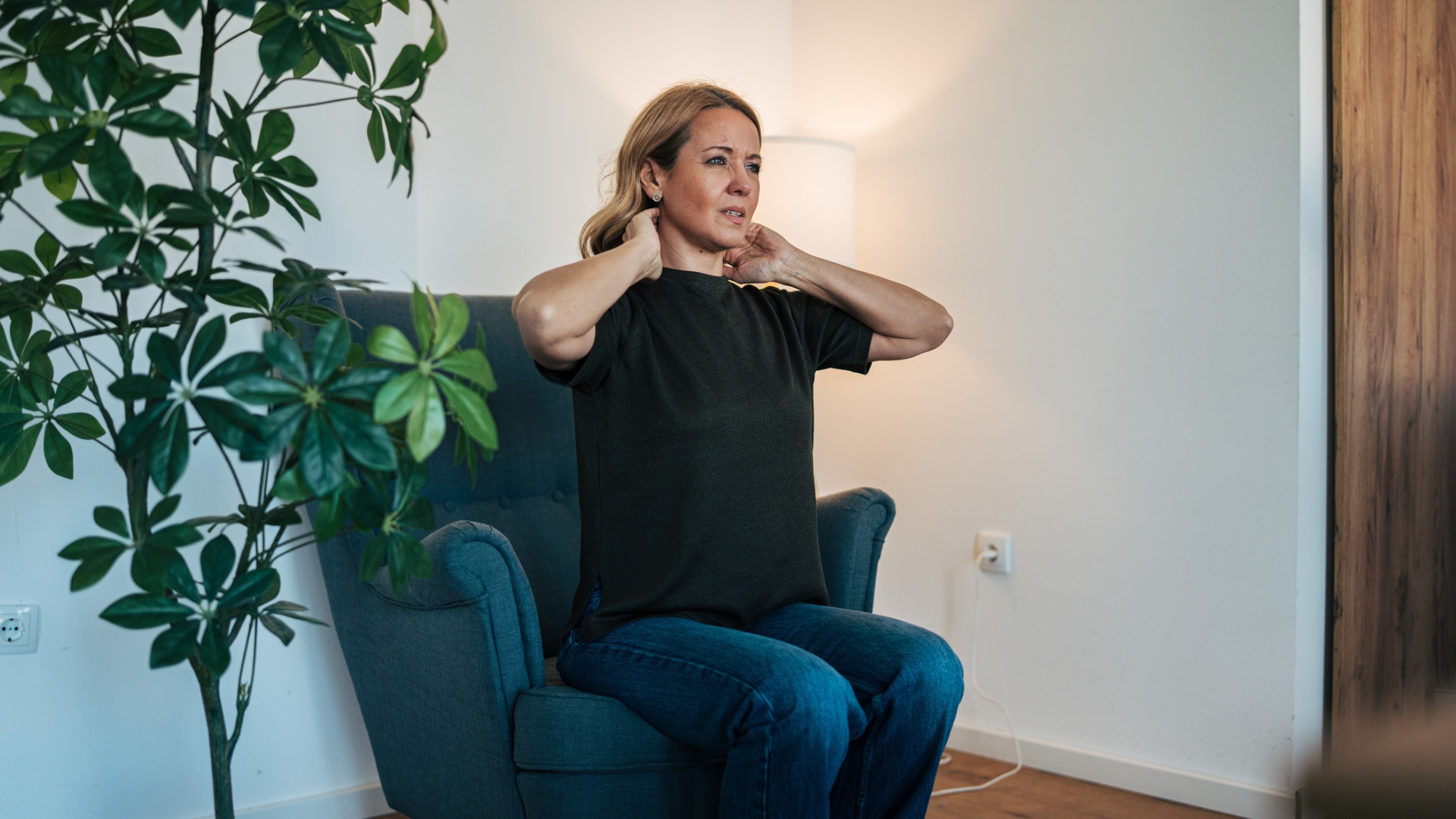 woman sitting on a velvet navy armchair looking off camera holding her neck. there's a plant to her side and a white wall behind her. 