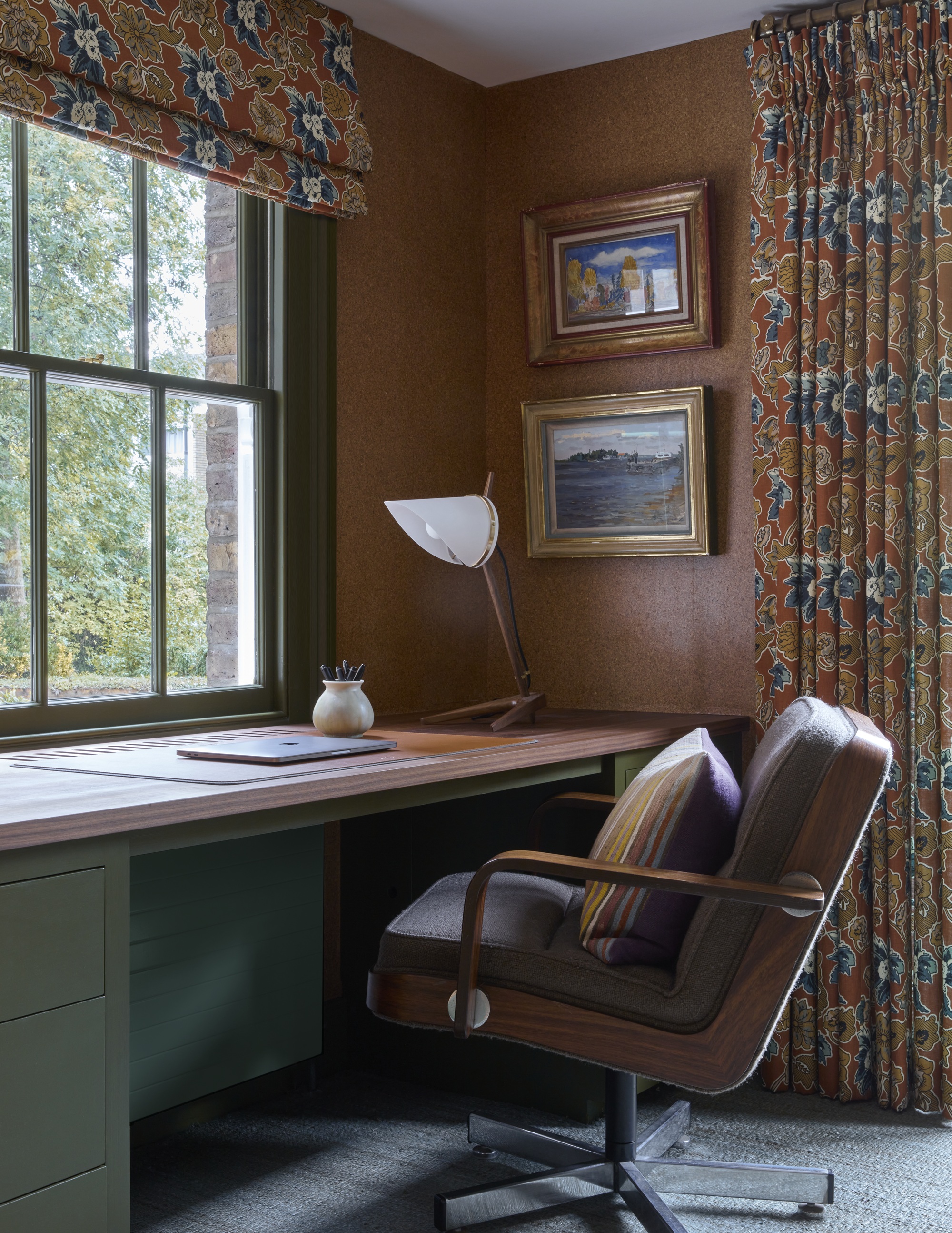 home office with walls clad in cork with patterned drapes and blinds, sage green desk with timber top, and artworks hung on the wall