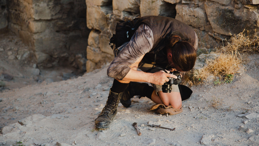 A man with long dark hair tied back in a ponytail kneels on his left knee on the ground. He aims a black camera at the ground. In the background, old walls and dirt.