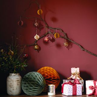 Red and golden Christmas baubles on branch in front of red wall, paper baubles and Christmas presents on wooden table