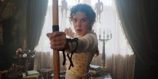 Enola Holmes (Millie Bobby Brown) pulls back the strong of an arrow.