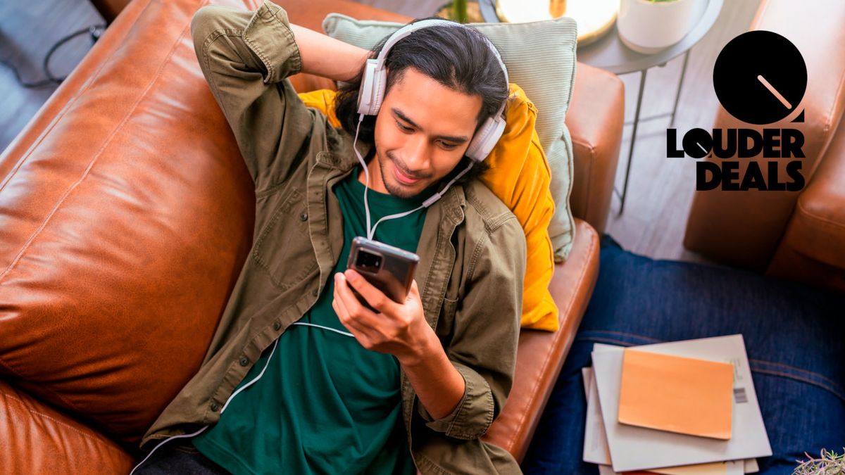 A young male lying on his back on a sofa, wearing headphones and holding a smartphone listening to music.