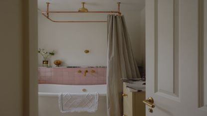 a simple yet pretty bathroom with a freestanding bath shown with pale pink tiles, gold shower fittings and a striped shower curtain
