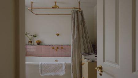a simple yet pretty bathroom with a freestanding bath shown with pale pink tiles, gold shower fittings and a striped shower curtain
