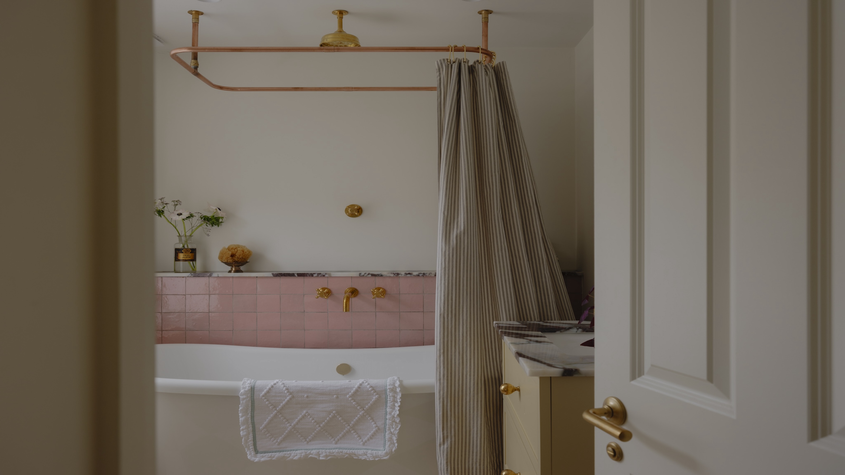 a simple yet pretty bathroom with a freestanding bath shown with pale pink tiles, gold shower fittings and a striped shower curtain