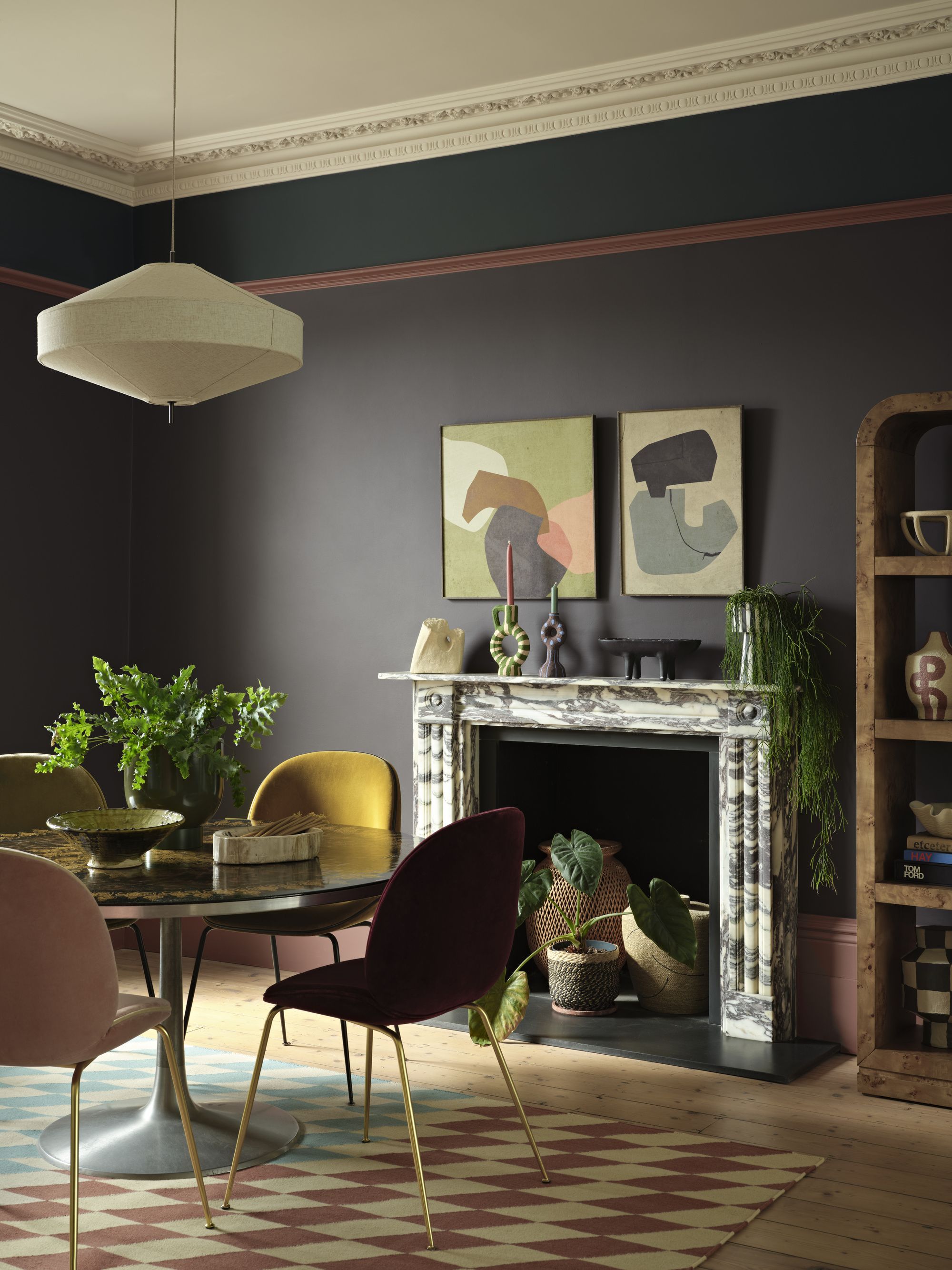 Image of a dining area in a room that is painted in a dark grayish-purple color with pink and teal trim. There is a marble fireplace with two paintings above it and a round dining table with jewel-toned seating.