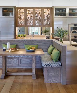 An L-shaped kitchen with a gray wooden dining nook with green throw pillows and a wooden table, silver countertops and cabinets, and a wooden floral accent panel