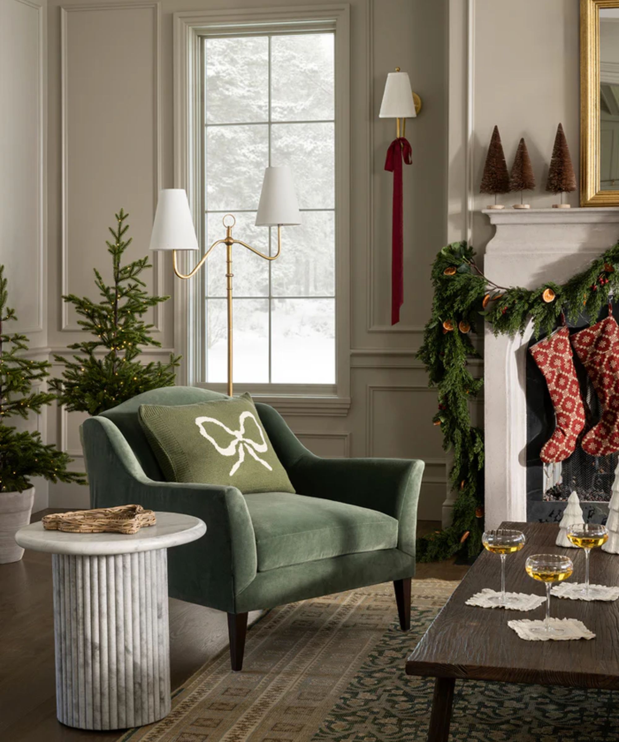 mcgee and co neutral living room with a white wooden fireplace styled with a foliage garland, dried oranges, red stockings, wire brush christmas trees and a large rug with a green armchair with a pillow with a bow on the front