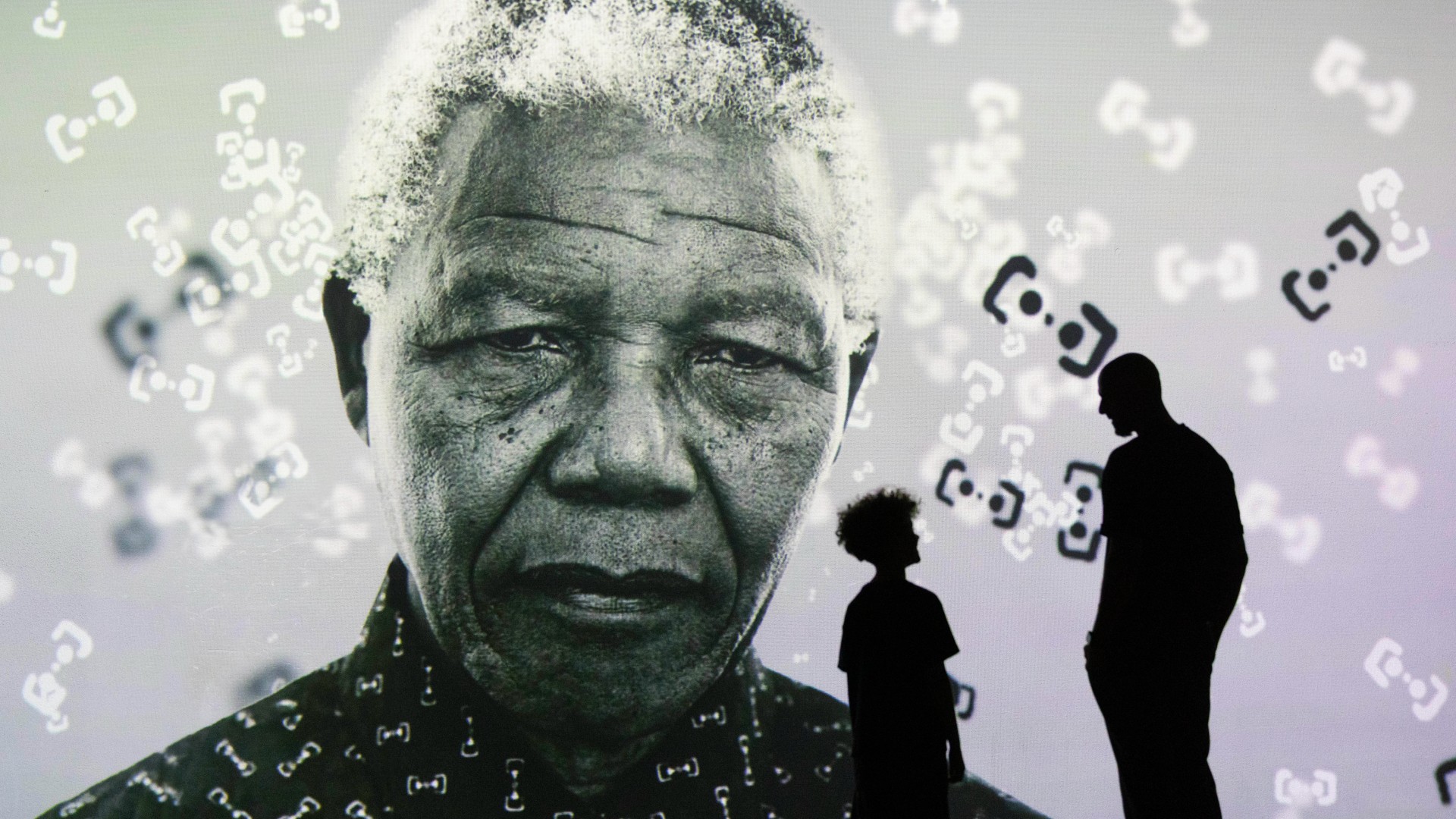 The silhouettes of an adult and a child stand before a vast black-and-white projection of Nelson Mandela's portrait at FRAMELESS London, with floating abstract symbols scattered across the image.