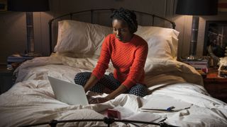 A woman in a red jumper sits on her bed looking up her mattress warranty on her laptop
