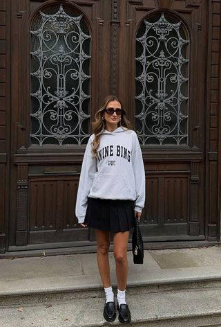 A woman standing in a doorway wearing a gray sweatshirt with a black pleated miniskirt, white socks, and black loafers.