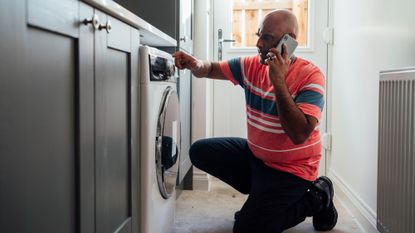 A man crouching down near his washing machine, calling an appliance repair service