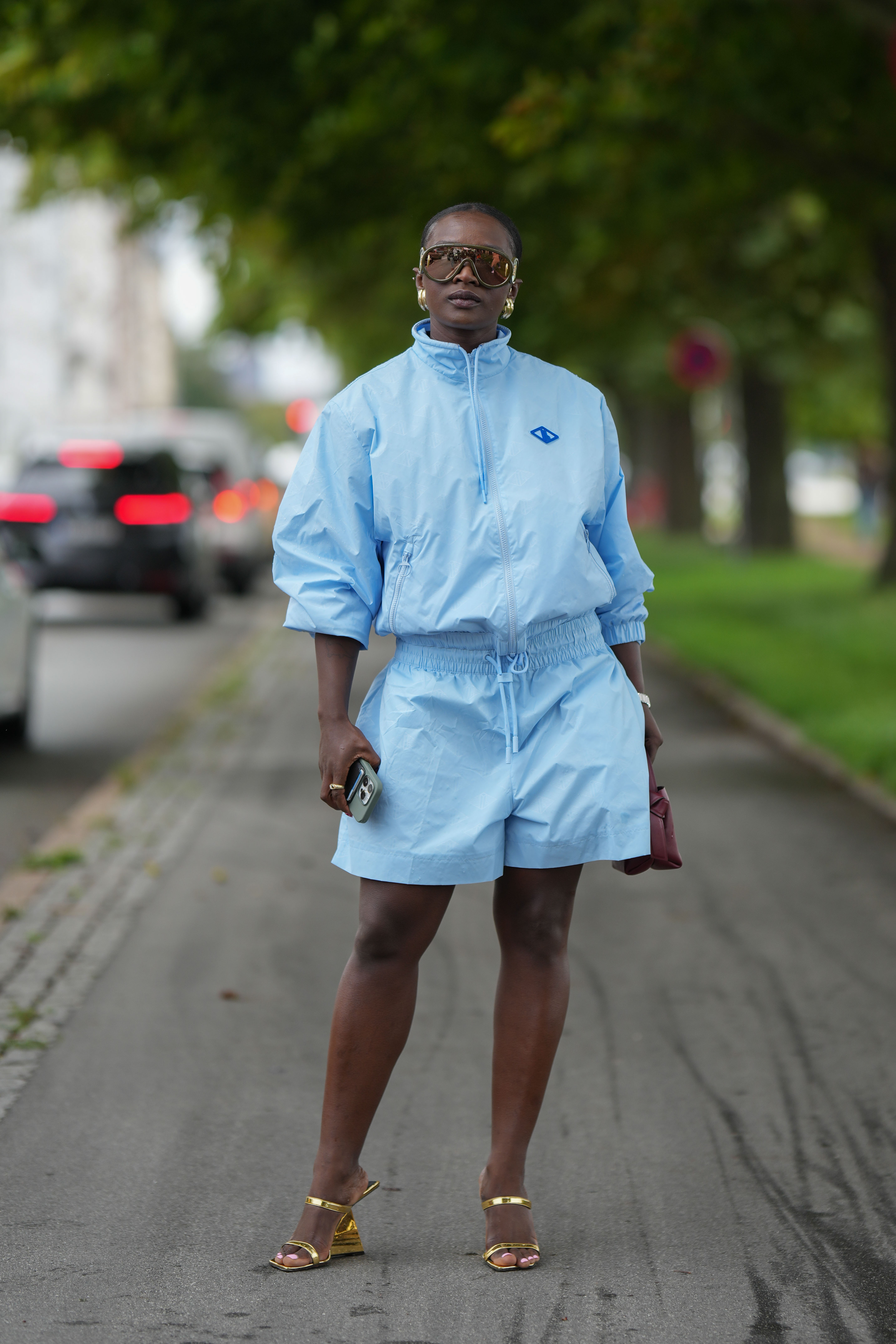 A guest wears a blue sport jacket, blue board shorts, a burgundy matte leather handbag, gold rings, gold shiny varnished leather strappy heels mules sandals on August 09, 2023 in Copenhagen, Denmark.