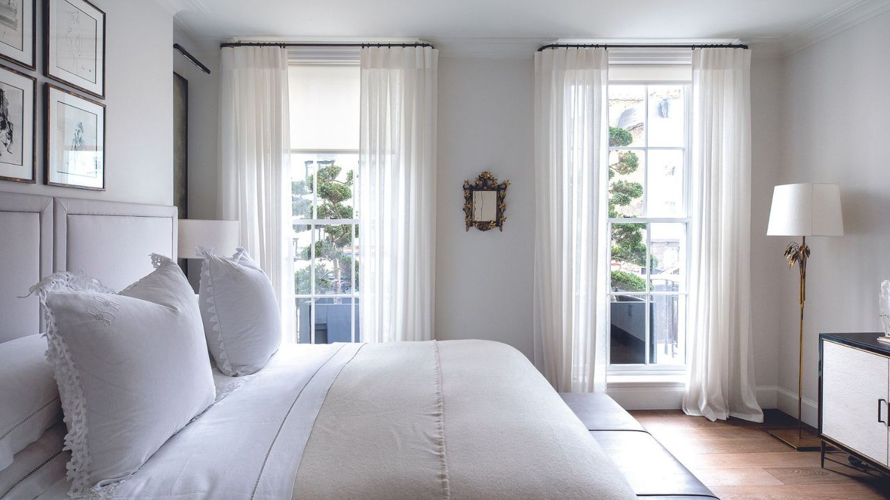 A cream bedroom with a white bed and sheer ivory linen curtains over floor to ceiling windows