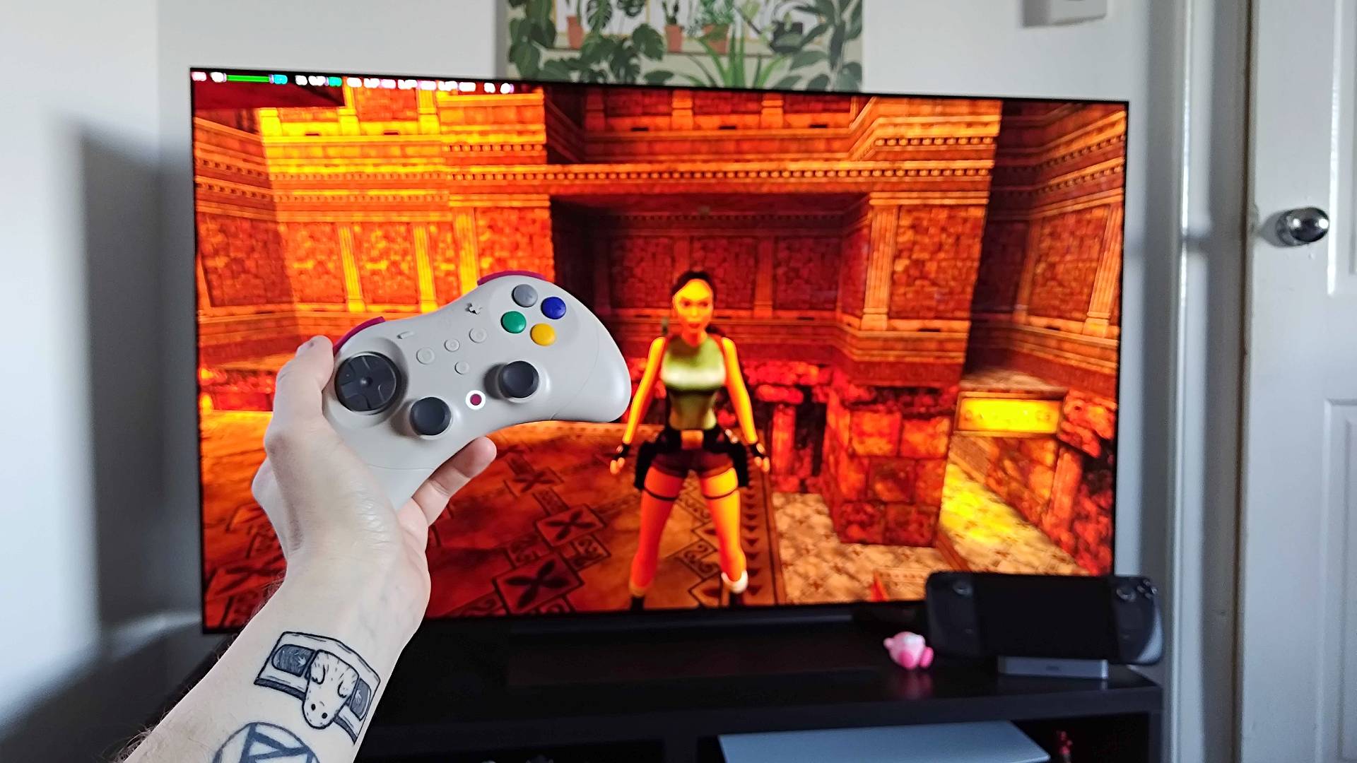 Hand holding Gulikit Elves 2 Pro controller in front of LG OLED TV with Tomb Raider gameplay in screen via docked Steam Deck.