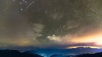 A breathtaking night sky filled with streaks of light from the Geminid meteor shower, set against a backdrop of twinkling stars and partially cloudy skies. Below, rugged mountain peaks are silhouetted, with a faint glow of city lights casting a soft illumination. The horizon features vibrant hues of orange and purple, blending into the dark, starry expanse above.