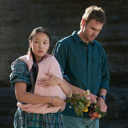 Yerin Ha as Mia and Charlie Vickers as Kieran walking with their baby and holding flowers in the survivors