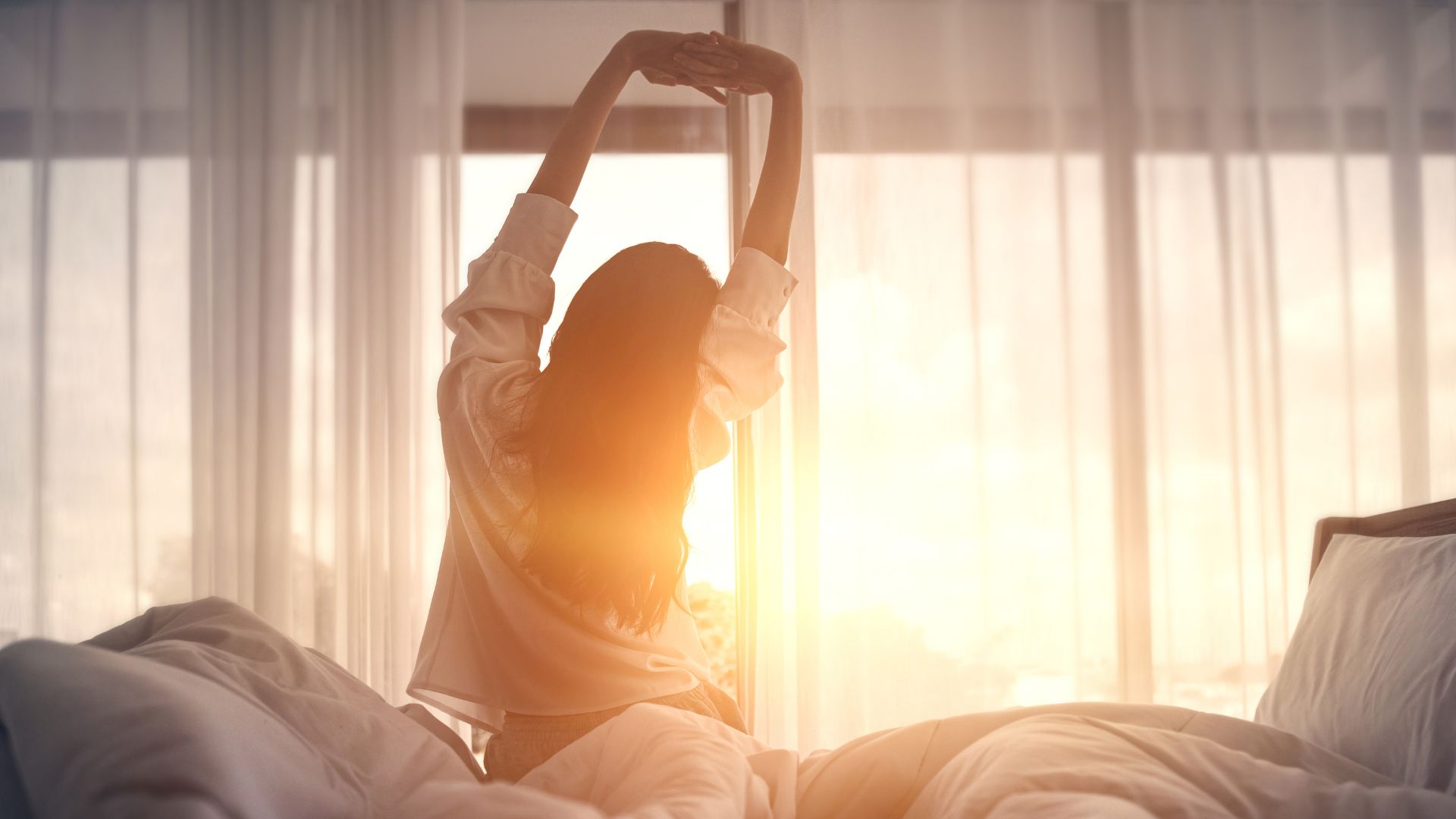 Woman facing away from the camera, sitting on the side of a bed and stretching in front of a window showing the sun rising.